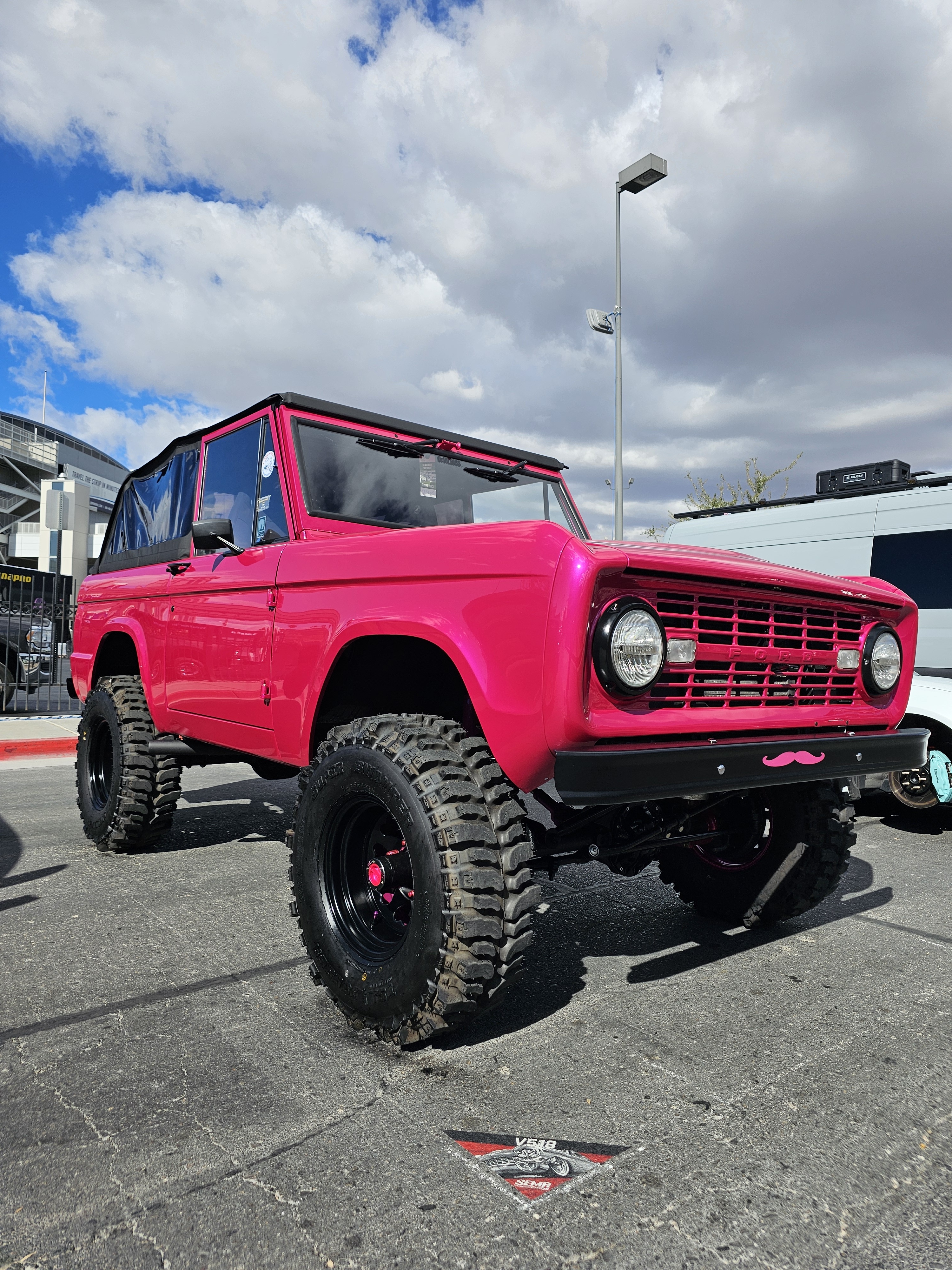 A bright pink Bronco with a pink mustache decal below the grille sits in a lot.